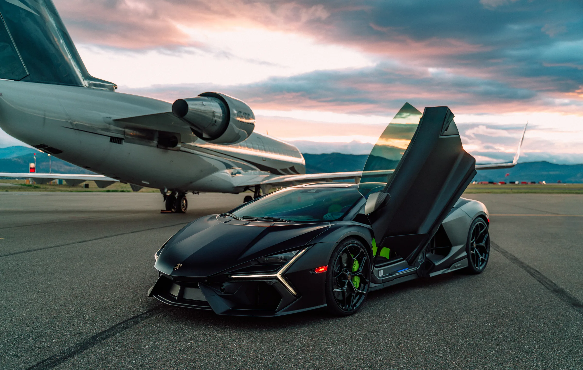 Matte black Lamborghini Revuelto with doors up beside a private jet on a Montana runway at dusk
